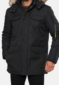 Sort parka-jakke med en hætte foret med pels. Har flere frontlommer og en lynlåslomme på ærmet. Glat stoftekstur og isoleret design.