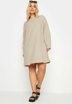 Vestido de suéter de canalé beige con un corte holgado y mangas largas. Presenta un dobladillo curvado y se combina con sandalias negras con tira en el tobillo.