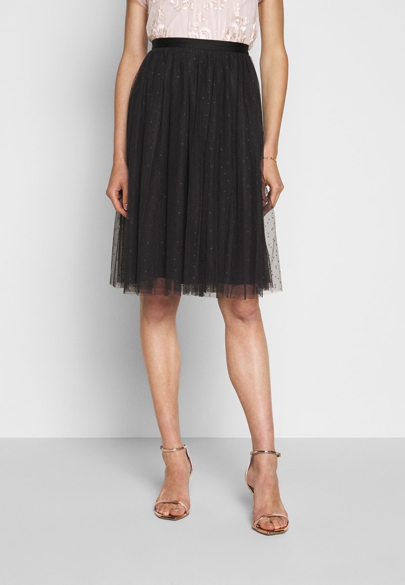 Femme portant une jupe en tulle noire mi-longue à petits pois, un haut floral rose clair et des talons nus à bride à la cheville, debout sur un sol gris.