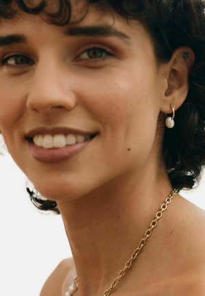 Uśmiechnięta osoba z krótkimi kręconymi włosami, nosząca złoty łańcuszek i złote kolczyki koła z białymi perłami, na jasnym tle.