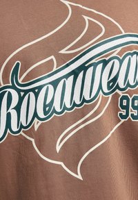 Brown fabric t-shirt featuring large white and teal "Rocawear 99" text with swirling accents and a textured surface.