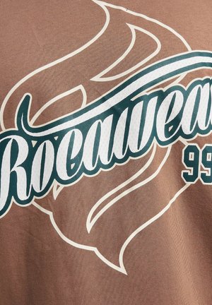 Brown fabric t-shirt featuring large white and teal "Rocawear 99" text with swirling accents and a textured surface.