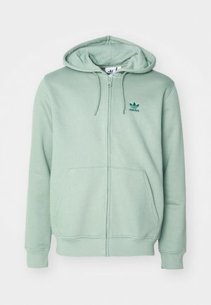 Jasnozielona bluza zapinana na zamek, z miękkiego materiału, z kapturem, kieszeniami z przodu i małym ciemnozielonym logo Adidas na lewej piersi.