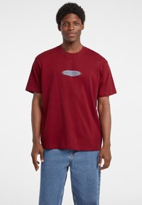 Burgundinės medvilninės trumpomis rankov�ėmis marškinėliai su apvaliu iškirpimu, priekyje su juodos ir baltos spalvos "GUESS JEANS" logotipo. 