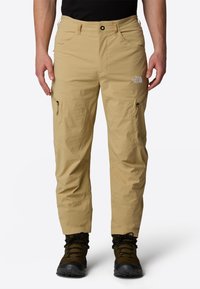 Beige waterproof pants with zippered pockets, adjustable waistband, and reinforced seams, paired with black hiking boots.