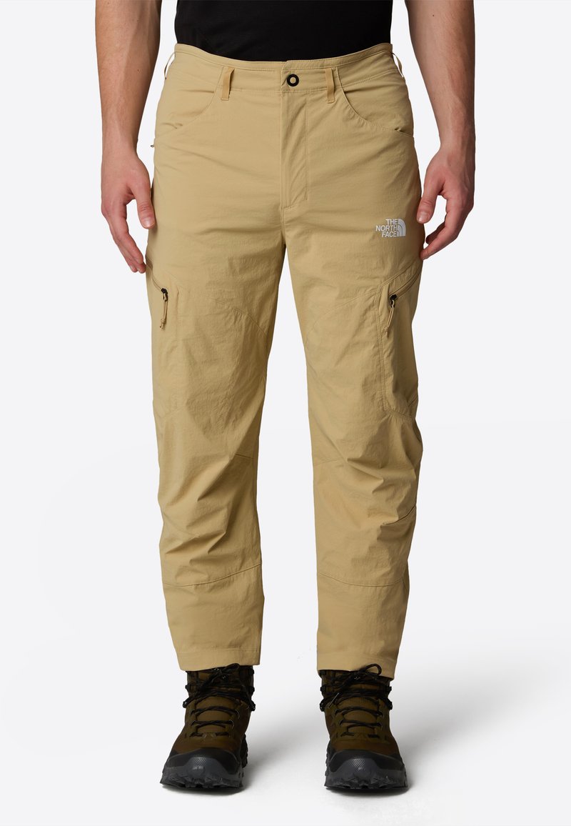 Beige waterproof pants with zippered pockets, adjustable waistband, and reinforced seams, paired with black hiking boots.