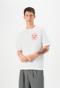 T-shirt en coton blanc avec un imprimé graphique rouge sur la poitrine gauche. Le modèle porte un pantalon plissé gris. Texture lisse et coupe décontractée.