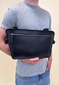Svart läderväska med en platt rektangulär form, med en silverfärgad dragkedja och justerbara remmar, hålls mot en grå skjorta.