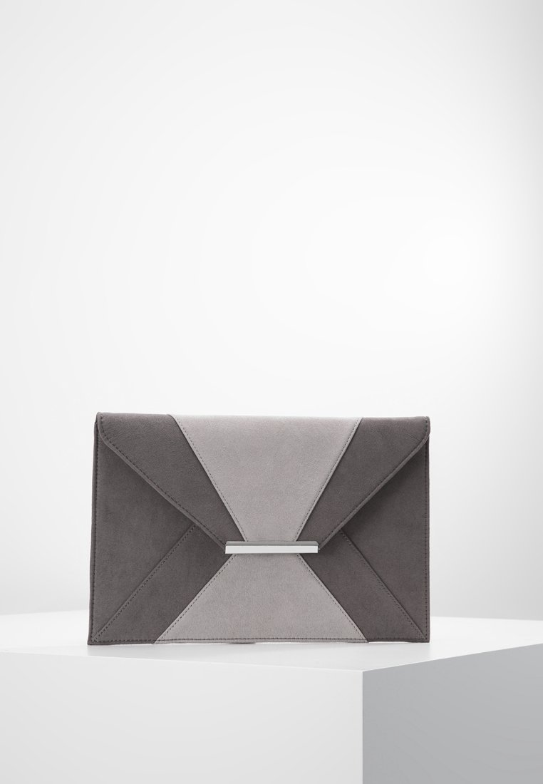 Pochette enveloppe en daim gris avec un design à panneaux géométriques et une fermeture rectangulaire argentée, posée sur un piédestal blanc sur fond clair.