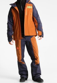 Veste et pantalon de ski en orange et bleu marine, fabriqués en matériau imperméable. Dotés de grandes poches, de fermetures éclair et d'un logo bien visible sur la poitrine.