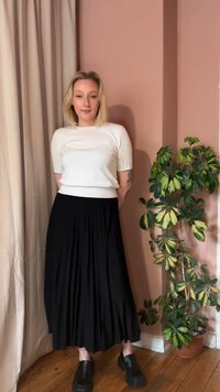 Žena plave kose stoji uz ružičasti zid noseći bijelu majicu kratkih rukava, crnu plisiranu suknju i crne platforme, pored saksijske biljke.