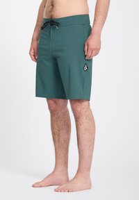 Homme portant un short de bain vert foncé arrivant aux genoux, avec un cordon noir et un logo sur la cuisse gauche, debout pieds nus devant un fond blanc.