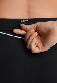 Black fitted leggings with a white accent at the waist. A hand is holding the waistband, showing a logo in white. Smooth texture.