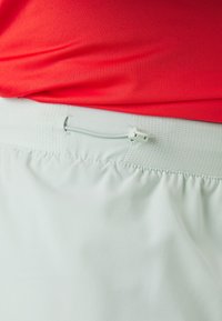 Pantalones cortos verde claro con una cinturilla texturizada, que presenta un cierre con cordón. La parte superior es una camiseta roja ajustada.