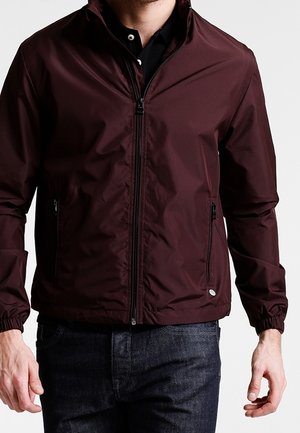 Mand iført en mørk bordeaux lynlåstrøje med lynlåslommer i siderne og elastiske ærmegab over en sort skjorte og mørke jeans.