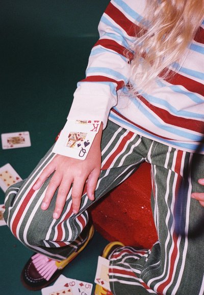 Gestreepte lange mouwen shirt in rood, blauw en wit. Groene verticaal gestreepte broek. Speelkaarten op een rode blok met vingers rustend op het been.