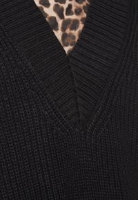 Svart texturerad stickad tröja med V-ringning, med leopardmönstrat tyg synligt under, vilket skapar kontrast.