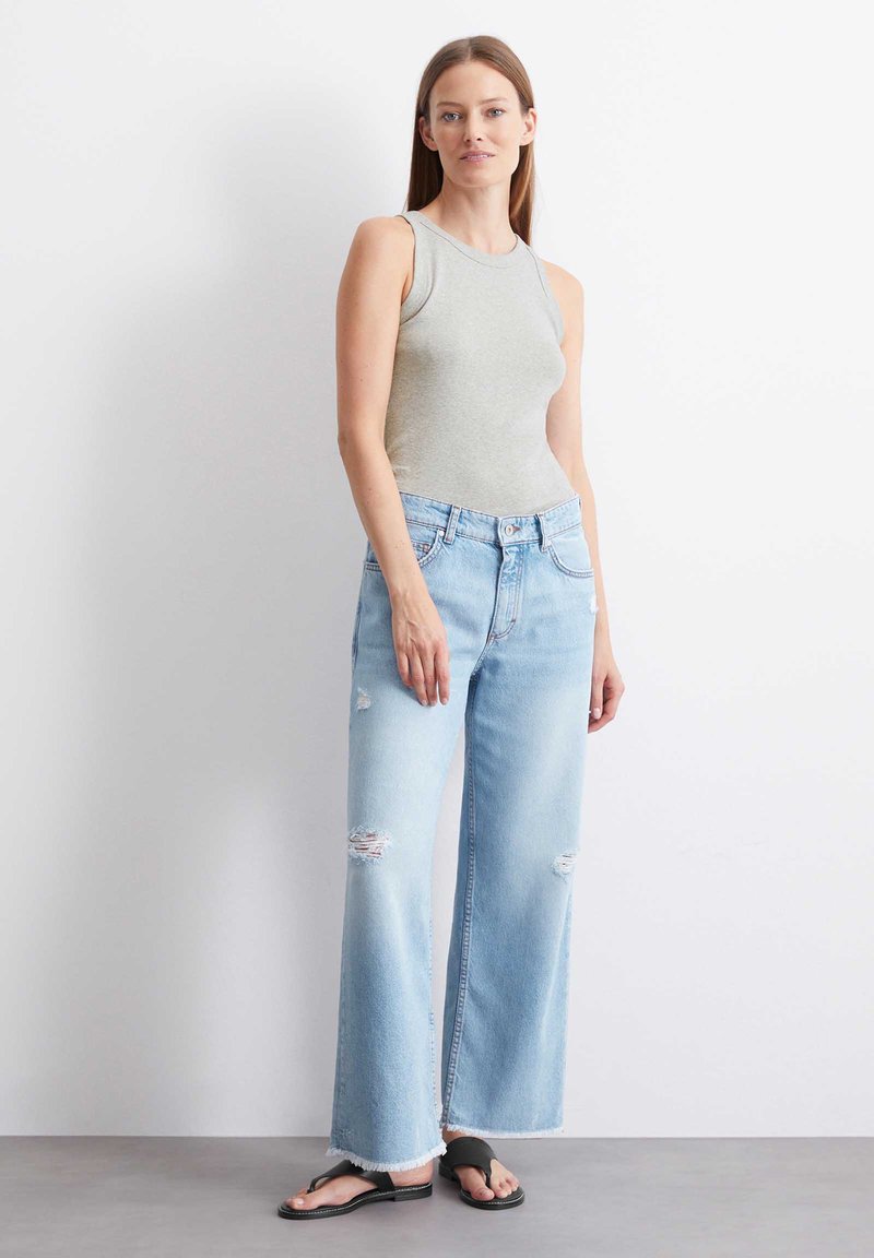 Jasnoszara koszulka na ramiączkach połączona z jasnoniebieskimi, zniszczonymi spodniami z szerokimi nogawkami. Dżinsy mają postrzępione końce nogawek i przetarcia dla dodatkowego efektu. Czarne klapki.