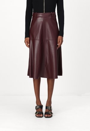 Vrouw die een donkere bruine leren midirok, een zwart shirt met lange mouwen en zwarte sandalen met grote gouden gespen draagt.