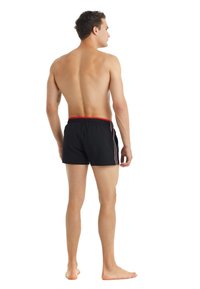 Costumi da bagno neri con elastico rosso in vita, strisce laterali e design aderente, indossati da una persona vista da dietro.
