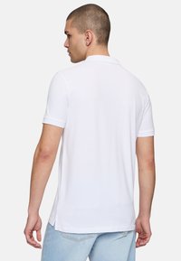 Valkoinen lyhythihainen polo paita, jossa on klassinen kaulus, valmistettu sileästä kankaasta. Siinä on sivuhalkiot ja rento leikkaus. Paita on yhdistetty farkkuliin.