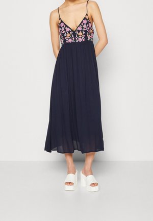 Navy blue maxi dress with spaghetti straps and a floral embroidered bodice, featuring a flowing skirt and white chunky platform heels.