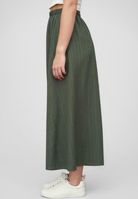 Lang, grønn, plisserte skjørt med elastisk midjebånd, laget av lett stoff, kombinert med hvite joggesko. Sidevisning vist.