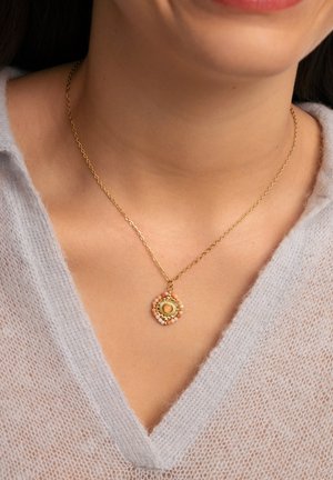 Collier en chaîne en or avec un pendentif rond présentant un cercle central en or, entouré de petites perles roses et blanches, sur un pull gris clair.