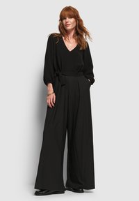 Combi-short noire avec jambes larges, décolleté en V plongeant et taille ceinturée. Présente des manches bouffantes et une texture douce. Portée avec des chaussures noires.
