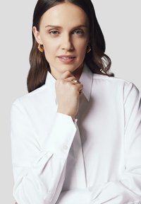 Biała koszula z długim rękawem i klasycznym kołnierzykiem, o gładkiej fakturze. Modelka nosi złote kolczyki w kształcie kół i srebrny pierścionek.