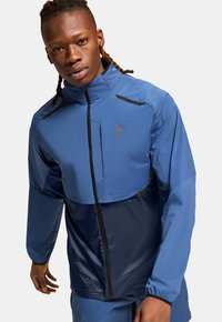 Blue athletic jacket featuring a quarter-zip design, contrasting navy panel, black accents, and a lightweight, water-resistant material.