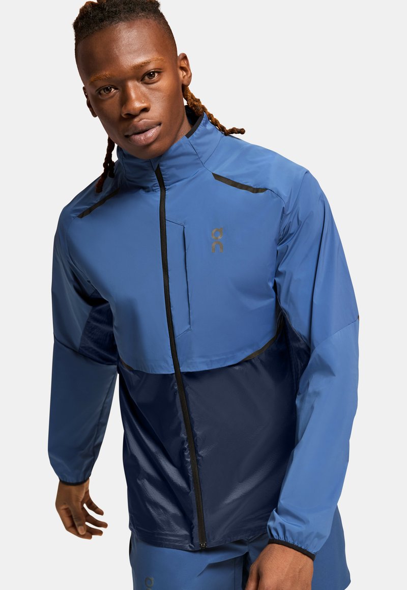 Blue athletic jacket featuring a quarter-zip design, contrasting navy panel, black accents, and a lightweight, water-resistant material.