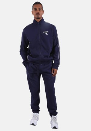 Tuta blu navy in tessuto morbido, con giacca con zip e dettagli bianchi e pantaloni abbinati. Scarpe da ginnastica sullo sfondo.