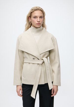 Femme blonde portant un manteau en laine beige ceinturé par-dessus un pull à col roulé assorti et un pantalon noir, debout devant un fond clair uni.