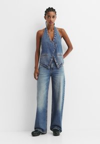 Denim obleka brez rokavov z ovratnikom halter, gumbi in dvema sprednjima žepoma, v kombinaciji s širokimi modrimi kavbojkami in črnimi leseni copati.