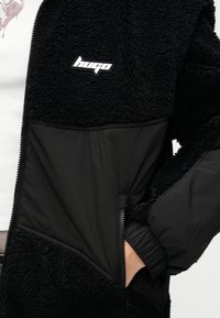 Μαύρο μπουφάν από φλις με αντίθετο λείο πάνελ, με λογότυπο, φερμουάρ και ελαστικά μανσέτα. Διακρίνεται υφασμάτινη υφή.