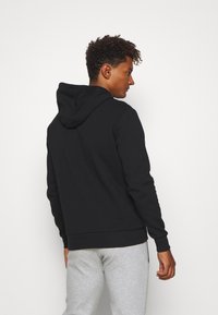 Schwarzer Kapuzenpullover mit einer glatten Textur. Verfügt über eine vordere Kängurutasche und gerippte Bündchen, getragen mit hellgrauen Jogginghosen.