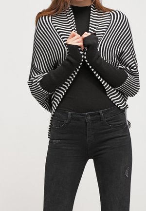 Frau trägt einen schwarz-weiß gestreiften Schal über einem schwarzen Langarmshirt und schwarzen, hoch taillierten Jeans mit dezenten Used-Details.