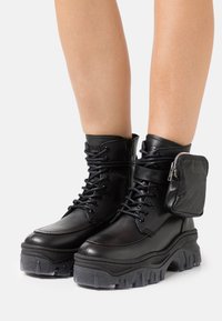 Bottes chelsea en cuir noir épais avec semelles crantées épaisses, lacets à l’avant, fermeture éclair latérale et petite pochette zippée attachée sur le côté extérieur.