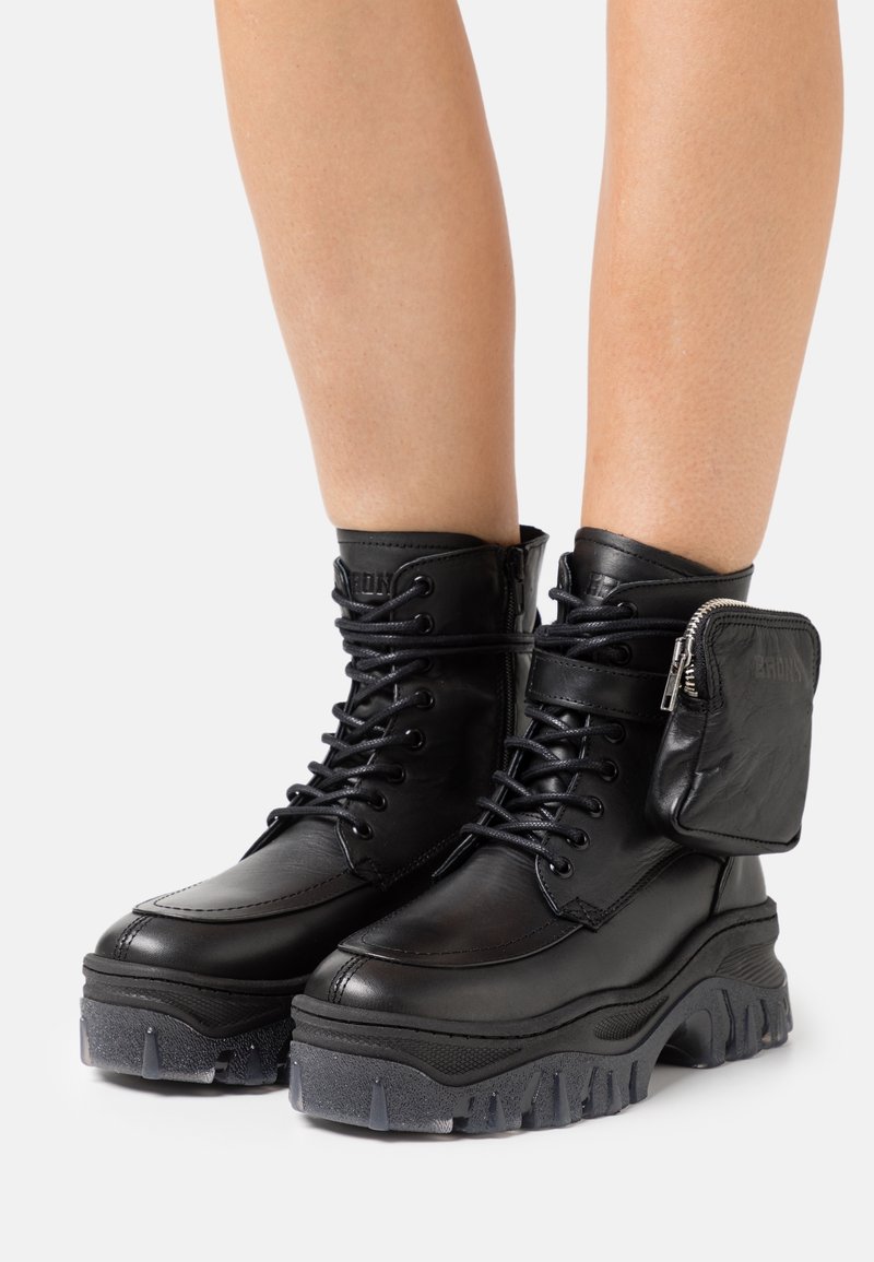 Bottes chelsea en cuir noir épais avec semelles crantées épaisses, lacets à l’avant, fermeture éclair latérale et petite pochette zippée attachée sur le côté extérieur.
