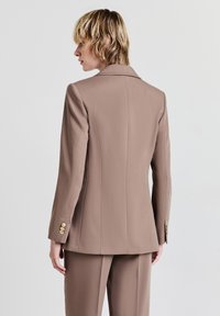 Blush-beiger, maßgeschneiderter Blazer aus strukturiertem Stoff mit klassischem Kragen, langen Ärmeln und goldfarbenen Knopfakzenten.