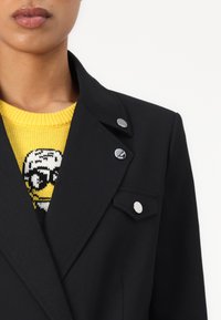 Blazer nero con colletto appuntito, dotato di tre bottoni argentati e una piccola tasca. Sotto, un maglione di maglia gialla con dettagli grafici.