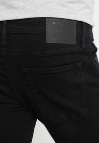 Vaqueros de mezclilla negros con una textura suave, corte ajustado y un distintivo parche de cuero negro en la cinturilla. Diseño clásico de bolsillo trasero.