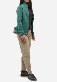Femme portant une veste verte zippée, un t-shirt noir, un pantalon beige et des chaussures de randonnée noires, se tenant légèrement de côté.