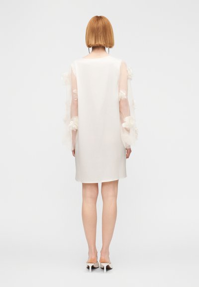 Άσπρο φόρεμα με διάφανα μανίκια διακοσμημένα με λουλουδένιες εφαρμογές, ίσια γραμμή, μήκος μέχρι το γόνατο, λείο ύφασμα, συνοδεύεται από άσπρα ψηλοτάκουνα παπούτσια.