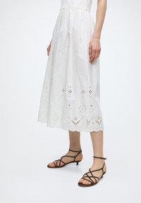 Γυναίκα που φοράει λευκό midi φόρεμα με κεντήματα και κοψίματα, και μαύρα σανδάλια με χαμηλό τακούνι και λουράκια, στέκεται μπροστά σε απλό φόντο.