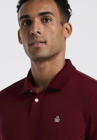 Bordeaux polo shirt met een kraag, drie knopen en een klein pinguïnlogo op de linkerborst. Gladde textuur en een getailleerd ontwerp.