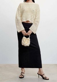 Häkeloberteil in Beige mit langen Ärmeln und offenem Muster, kombiniert mit einem schwarzen Maxirock. Akzentuiert durch eine kleine Muschel-Handtasche und schwarze Riemchenpumps.