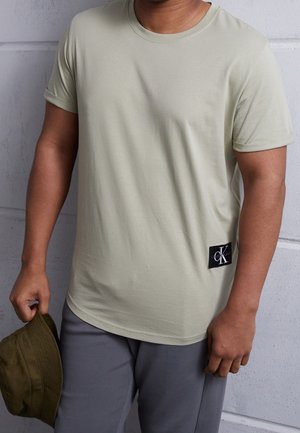 Jasnozielona koszulka z okrągłym dekoltem, krótkimi podwiniętymi rękawami oraz czarną metką z białym napisem po lewej stronie.