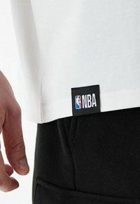 Camiseta blanca de manga corta, con una etiqueta negra de la NBA en el dobladillo; combinada con pantalones negros, que presentan un diseño sencillo.